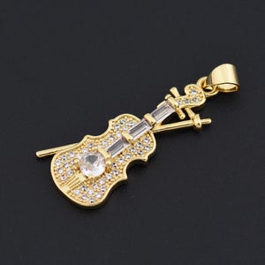 May include: A gold-colored violin pendant with a clear, round gemstone at the center and smaller clear stones throughout. The pendant includes a bow and a loop for a chain. The background is black.