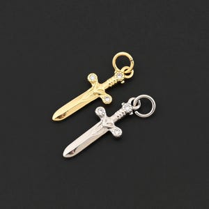 Puede incluir: Dos pequeños colgantes de espada, uno dorado y otro plateado, sobre un fondo negro. Cada colgante presenta una empuñadura detallada con detalles de piedras preciosas transparentes y un pequeño lazo para sujetar. Las espadas miden unos 2,5 cm de largo.