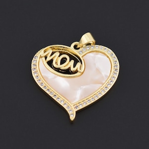 Könnte beinhalten: Goldfarbener Herzanhänger mit dem Wort „MOM“ in Schreibschrift. Der Anhänger hat einen Perlmutt-Mittelteil, umgeben von einem Rand aus klaren Kristallen. Eine kleine Schlaufe oben ermöglicht das Anbringen einer Kette.