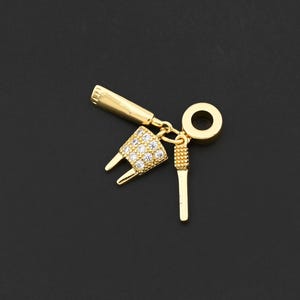 以下が含まれることがあります： 歯をテーマにした要素が特徴のゴールドカラーのチャーム。チャームには、小さな透明な石がちりばめられた歯の形をしたデザイン、歯科用ツール、小さなリングが含まれています。チャームは暗い背景に配置されています。