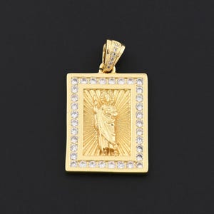 May include: Gold-colored rectangular pendant with a religious figure in the center, surrounded by a border of small, clear stones. The pendant has a loop at the top for a chain. The background is black.