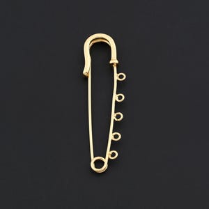 May include: A gold-colored safety pin with a series of small loops along one side. The pin is set against a black background, highlighting its bright metallic finish. The pin's design is simple yet elegant.