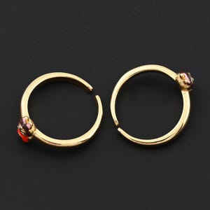 May include: Two gold-colored hoop earrings with a small, colorful gemstone accent. The earrings are open-ended and feature a gold finish. The gemstones have red, purple, and pink hues. The earrings are displayed on a black background.