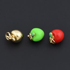 May include: Three apple-shaped charms in gold, green, and red. The gold charm is a smooth sphere. The green and red charms have a small green leaf and gold stem. The charms are on a black background.