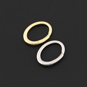 May include: Two oval metal clasps, one gold and one silver, are shown against a black backdrop. Each clasp is closed, with a small hinge visible. The gold clasp is positioned above the silver clasp. The clasps are likely used for jewellery or accessories.