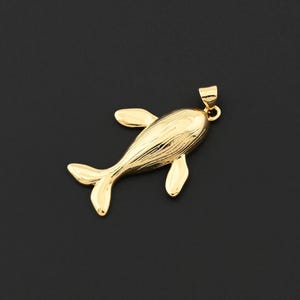 Può includere: Un ciondolo a forma di balena color oro con superficie strutturata. Il ciondolo ha un anello per una catena. La balena ha pinne e coda. Lo sfondo è nero.
