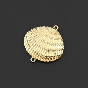 May include: A gold-coloured seashell charm with a textured surface. The charm has two small loops for attaching to jewellery. The seashell design is detailed with ridges and lines, giving it a realistic appearance. The charm is set against a dark background.