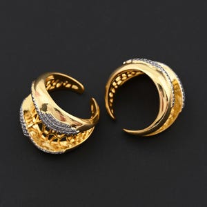 Könnte beinhalten: Zwei goldfarbene Creolen mit strukturiertem Design. Jeder Ohrring hat eine gebogene Form mit einem Band aus kleinen, funkelnden Steinen. Die Ohrringe sind vor einem dunklen Hintergrund platziert.