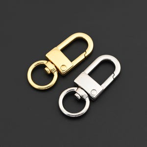 May include: Two metal swivel clasps, one gold-toned and one silver-toned, are displayed against a dark background. Each clasp features a rectangular base with a curved top and a circular ring for attachment.