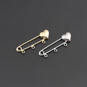 May include: Two safety pin style brooches, one in a gold tone and one in a silver tone. Each brooch features a smooth heart-shaped top and two small circular loops attached to the bottom bar for hanging additional charms or decorative elements.