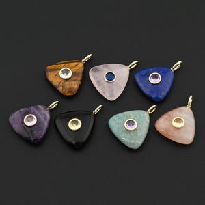 May include: Seven gemstone pendants in various colors, including tiger's eye, rose quartz, and amethyst. Each triangular pendant has a gold-colored bail and a small, round gemstone in the center. The pendants are displayed against a black background.