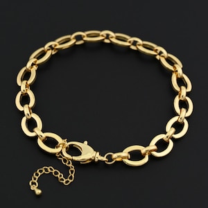 Puede incluir: Un brazalete de cadena dorada con eslabones ovalados. El brazalete tiene un cierre de mosquetón y una cadena ajustable. La joya se muestra sobre un fondo oscuro.