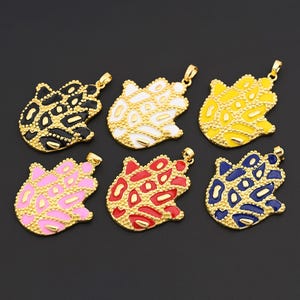 Peut inclure: Six pendentifs en métal doré en forme de grenouille. Chaque pendentif est rempli d'émail de couleur différente : noir, blanc, jaune, rose, rouge et bleu. Les pendentifs sont sur un fond sombre.