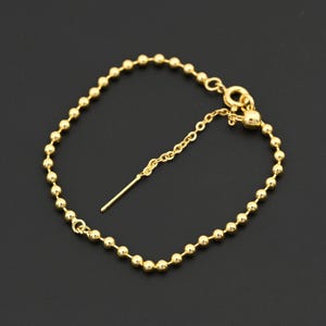May include: A gold-colored bracelet featuring a delicate chain of small, round beads. The bracelet has a sliding clasp for adjustable sizing and a small, dangling bar. The jewelry is displayed against a dark background.