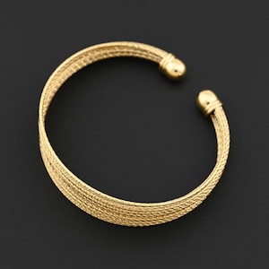 Peut inclure: Un bracelet manchette doré avec un motif tissé texturé. Le bracelet a des embouts arrondis et pleins. La conception ouverte permet de le porter facilement. Le bijou est posé sur un fond sombre.
