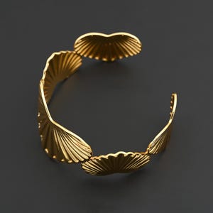 May include: A gold-colored cuff bracelet with a fan-like design. The bracelet features multiple stylized fan shapes, creating an open, adjustable design. The metal has a polished finish, reflecting light against a dark background.