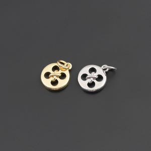 May include: Two circular metal charms shaped like four-hole buttons, one in a polished gold tone and one in a polished silver tone. Each charm features a central cross-stitch detail and a small jump ring attached to the top for hanging purposes.