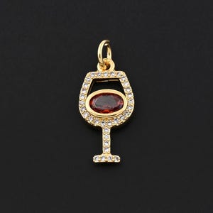 May include: A gold-colored pendant in the shape of a wine glass. The glass contains a red oval-shaped stone, surrounded by small clear stones. The stem and base of the glass are also adorned with clear stones. The pendant hangs from a small loop.