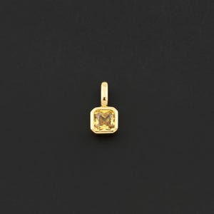 May include: A gold pendant with a square, yellow gemstone. The pendant has a small loop at the top for a chain. The gemstone is a light yellow color and is set in a gold bezel. The background is black.