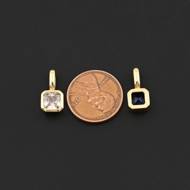 May include: Two gold-colored square pendants with gemstones, one clear and one dark blue, next to a penny. The pendants have a small loop at the top for hanging. The penny is dated 1976.