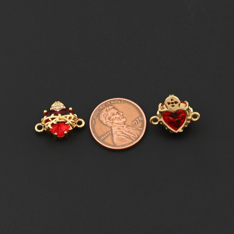 May include: Two gold-colored heart-shaped charms with red gemstone centers and small loops for attaching to jewelry. One charm has a crown design with small clear stones. A penny is next to the charms.