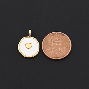 Puede incluir: Un colgante dorado con un centro de esmalte blanco y una forma de coraz&oacute;n dorada. El colgante est&aacute; al lado de un centavo para comparar el tama&ntilde;o. El colgante tiene un peque&ntilde;o lazo en la parte superior para una cadena.