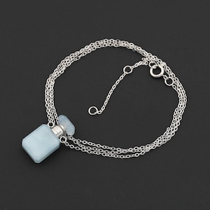 May include: A silver necklace featuring a light blue perfume bottle pendant. The pendant has a rectangular shape with a matching cap. The necklace chain is delicate and has a lobster clasp closure. The background is solid black.