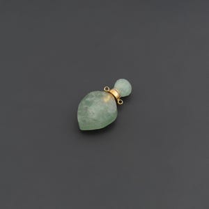 May include: A small, faceted, pale green stone perfume bottle pendant with a gold-toned metal cap. The cap features two small circular loops on opposite sides, allowing it to be attached to a chain or cord for use as a decorative jewelry piece.