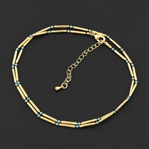 May include: Gold-colored anklet with a double-strand design. The anklet features small, light blue beads interspersed along the gold bars. The adjustable chain allows for a comfortable fit. The anklet is displayed against a black background.