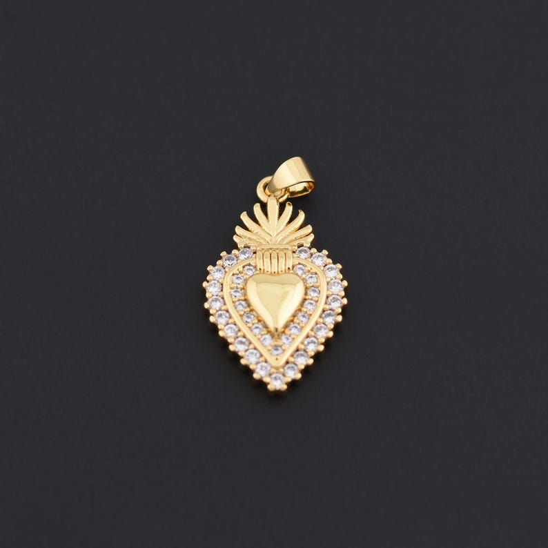 May include: A gold-colored pendant in the shape of a heart, adorned with clear crystals around the edges. The pendant has a bail for attaching to a chain. The design includes a heart shape in the center and a decorative top.