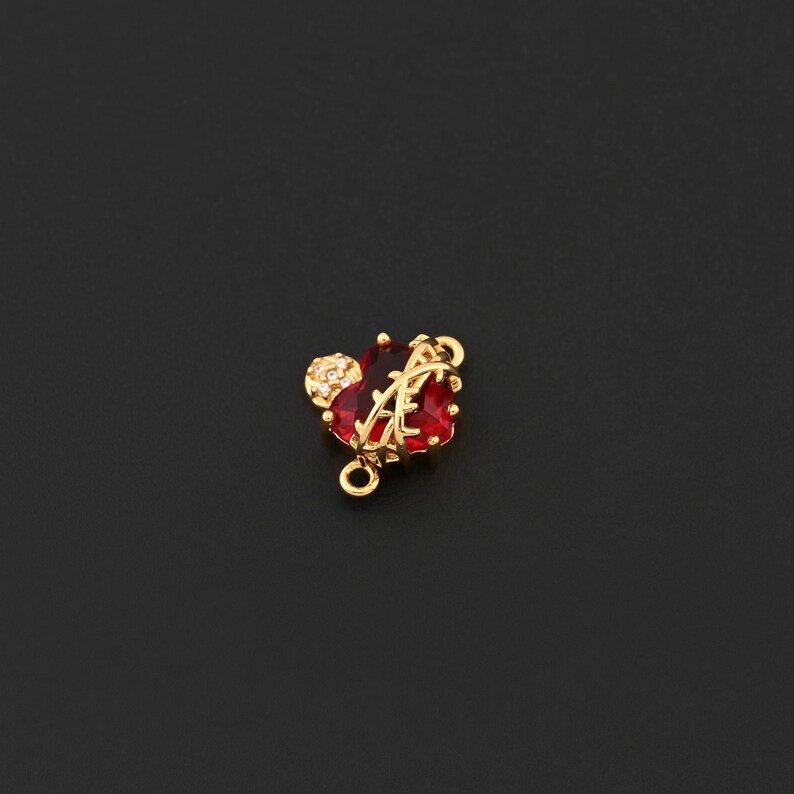 May include: A gold-colored charm with a red heart-shaped gemstone and a small, round, crystal-studded accent. The charm has a loop for attaching to jewelry and is set against a dark background.
