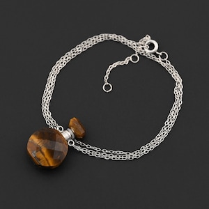 Puede incluir: Un collar de plata con un colgante facetado y redondo de ojo de tigre y un tapón a juego. El colgante cuelga de una delicada cadena de tres hilos. El collar se muestra sobre un fondo negro.