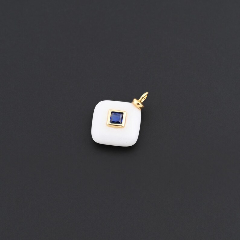May include: A white square pendant with a gold bail and a square, dark blue gemstone set in a gold frame. The pendant is set against a dark grey background.