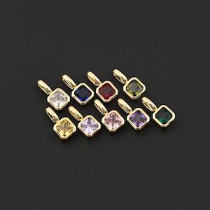 May include: A collection of ten gold-colored pendants, each featuring a square-cut gemstone in a different color. Gemstone colors include clear, blue, red, green, yellow, purple, and pink. The pendants are arranged on a black surface.