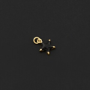 May include: A small, black star charm with gold-colored accents. The charm has a loop for attaching to a necklace or bracelet. The star's points are gold, and the charm is set against a dark background.