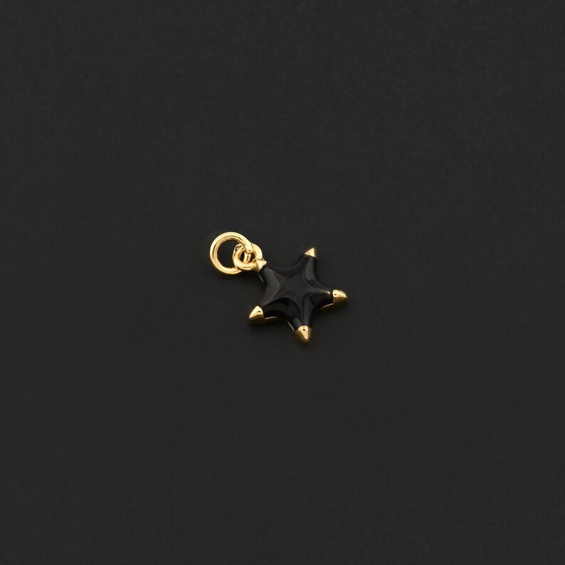 May include: A small, black star charm with gold-colored accents. The charm has a loop for attaching to a necklace or bracelet. The star's points are gold, and the charm is set against a dark background.