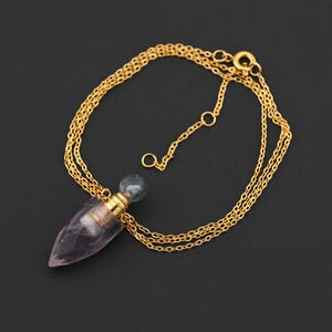 May include: A gold-colored necklace with a pendant. The pendant is a light purple, crystal-like, teardrop shape with a gold-colored cap and a small, clear, round top. The necklace has a triple-strand chain and a clasp.