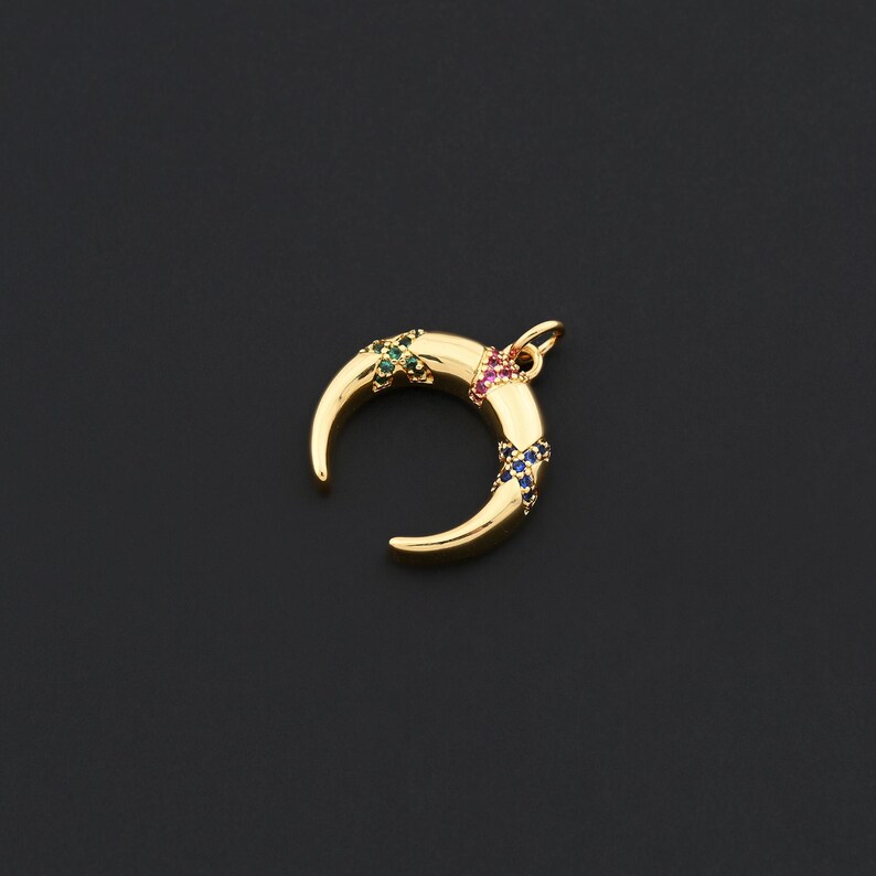 K&ouml;nnte beinhalten: Ein goldfarbener Halbmond-Anh&auml;nger mit bunten Edelsteinen. Der Anh&auml;nger hat eine gebogene Form mit gr&uuml;nen, rosa und blauen Edelsteinen. Der Hintergrund ist einfarbig schwarz.