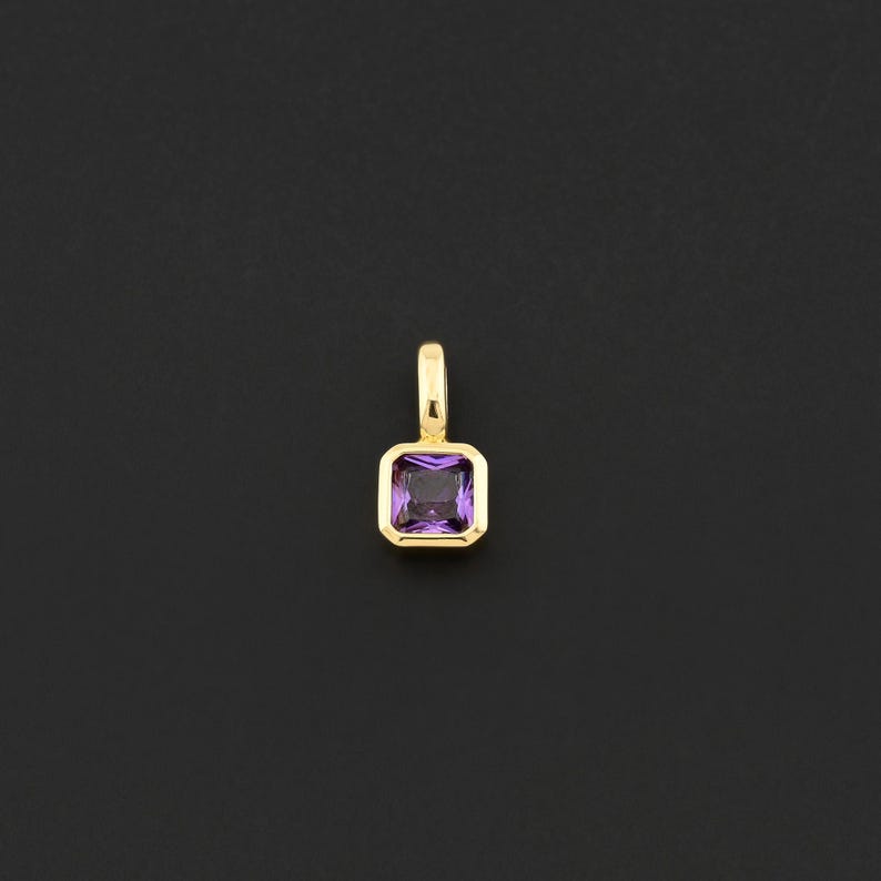 May include: A gold pendant with a square, purple gemstone. The gemstone is set in a gold bezel and has a rectangular shape. The pendant has a small loop at the top for a chain.