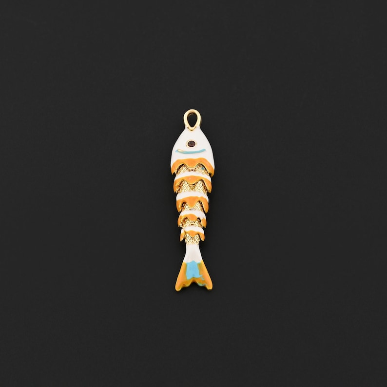 May include: A gold-colored fish pendant with white, orange, and blue enamel detailing. The fish-shaped charm has a loop at the top for hanging and is set against a black background. The pendant is likely for jewelry making.