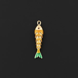 May include: A gold-colored fish pendant with a red gem eye and a green and yellow tail. The fish has a textured scale pattern and a loop at the top for hanging. The pendant is set against a black background.