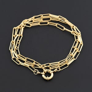 May include: A gold-colored necklace with a paperclip chain design. The necklace features multiple strands of rectangular links connected by a circular clasp. The jewelry is displayed against a dark background.