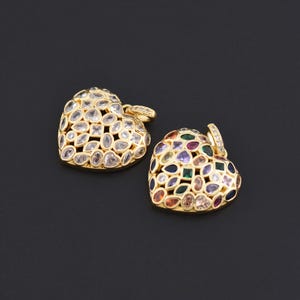 Peut inclure: Deux pendentifs en forme de cœur avec des montures dorées. L'un est recouvert de pierres claires et facettées, l'autre présente diverses pierres précieuses colorées. Les deux ont de petites boucles pour une chaîne, sur fond noir.