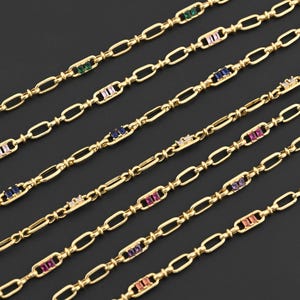 May include: Gold-colored chain necklaces with colorful rectangular gemstones. The gemstones are green, pink, blue, white, purple, and orange. The chains have an oval link design and are displayed on a black background.