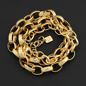 Puede incluir: Un collar de cadena dorado con eslabones texturizados y una pequeña etiqueta que dice "Good Luck". El collar se muestra sobre un fondo negro, resaltando su apariencia brillante y metálica. La cadena está hecha de múltiples eslabones ovalados.