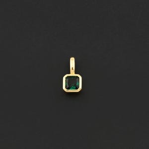 May include: A gold pendant with a square, emerald-cut green gemstone. The gemstone is set in a gold bezel and has a small gold loop at the top for a chain. The pendant is displayed against a black background.