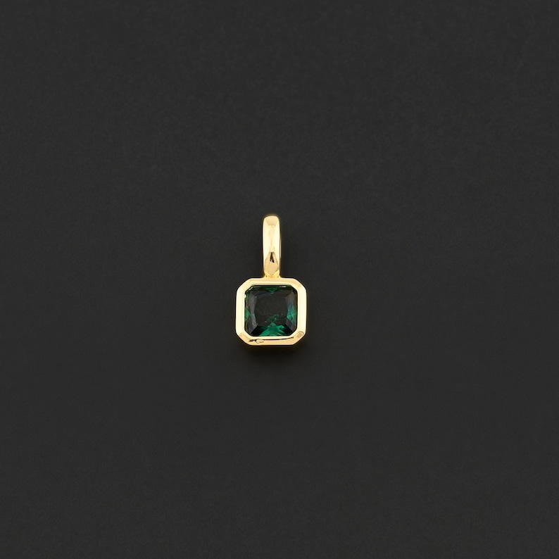 May include: A gold pendant with a square, emerald-cut green gemstone. The gemstone is set in a gold bezel and has a small gold loop at the top for a chain. The pendant is displayed against a black background.