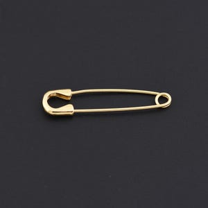 Puede incluir: Un imperdible dorado sobre un fondo negro. El imperdible tiene un diseño clásico, con una parte superior curvada y un cierre circular. El imperdible está hecho de un metal brillante y reflectante.