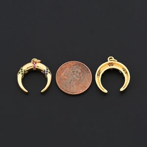 K&ouml;nnte beinhalten: Zwei goldfarbene Halbmond-Anh&auml;nger mit bunten Edelsteinen, neben einem Penny. Ein Anh&auml;nger hat gr&uuml;ne, rosa und blaue Edelsteine. Der andere hat rote Edelsteine. Die Anh&auml;nger befinden sich auf schwarzem Hintergrund.