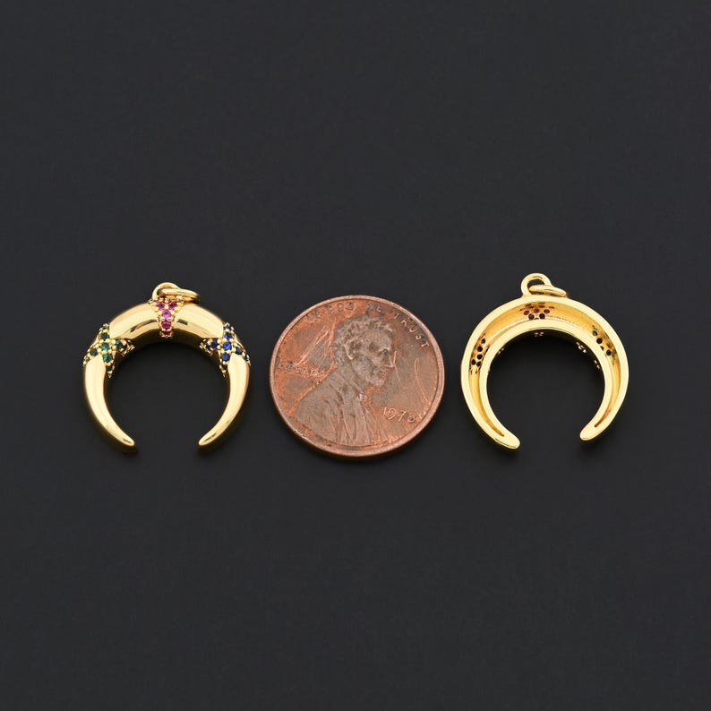 K&ouml;nnte beinhalten: Zwei goldfarbene Halbmond-Anh&auml;nger mit bunten Edelsteinen, neben einem Penny. Ein Anh&auml;nger hat gr&uuml;ne, rosa und blaue Edelsteine. Der andere hat rote Edelsteine. Die Anh&auml;nger befinden sich auf schwarzem Hintergrund.